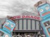 Composite image of the Cow Palace and parking log under a turbulent grey sky while blue toy robots approach the viewer