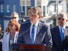 Short man in blue suit and skinny blue tie speaking at a lectern outdoors. Behind him are some of his supporters