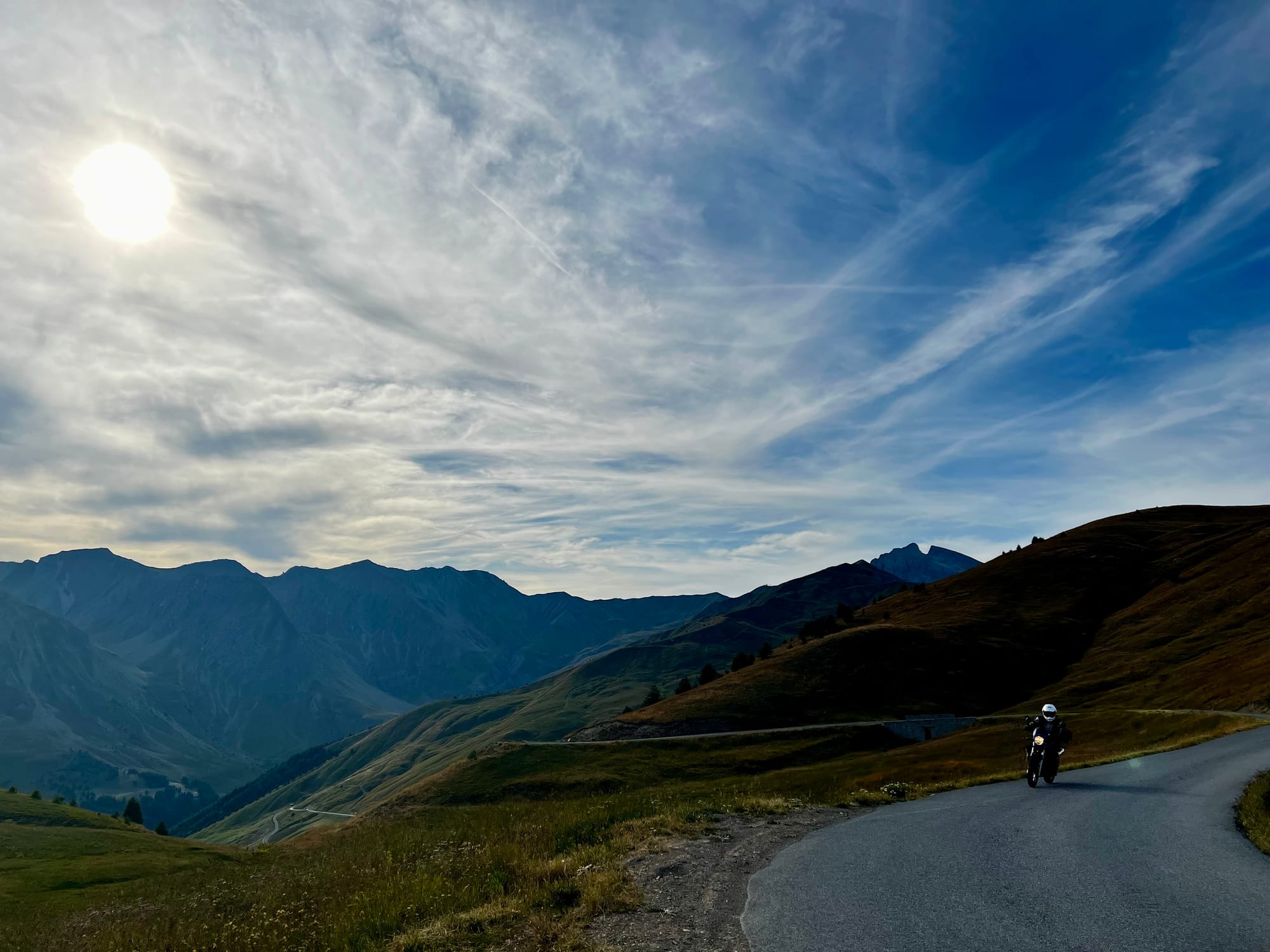 GPT  Das Bild zeigt eine kurvenreiche Bergstraße, die sich durch eine alpine Landschaft schlängelt. Im Vordergrund sieht man einen Motorradfahrer, der in voller Fahrerausrüstung auf einem Motorrad sitzt und die Straße entlangfährt. Die umliegenden Hänge sind mit spärlicher Vegetation bedeckt, was auf die Höhe und das rauere Klima hindeutet. Im Hintergrund erheben sich beeindruckende Berge, die unter einem weiten, mit zarten Wolken bedeckten Himmel liegen. Die Sonne scheint hell und steht hoch am Himmel, wobei ihre Strahlen die gesamte Szene in ein weiches Licht tauchen. Das Foto strahlt ein Gefühl von Abenteuer und Freiheit aus, das oft mit Motorradtouren in solch atemberaubenden Landschaften verbunden wird.