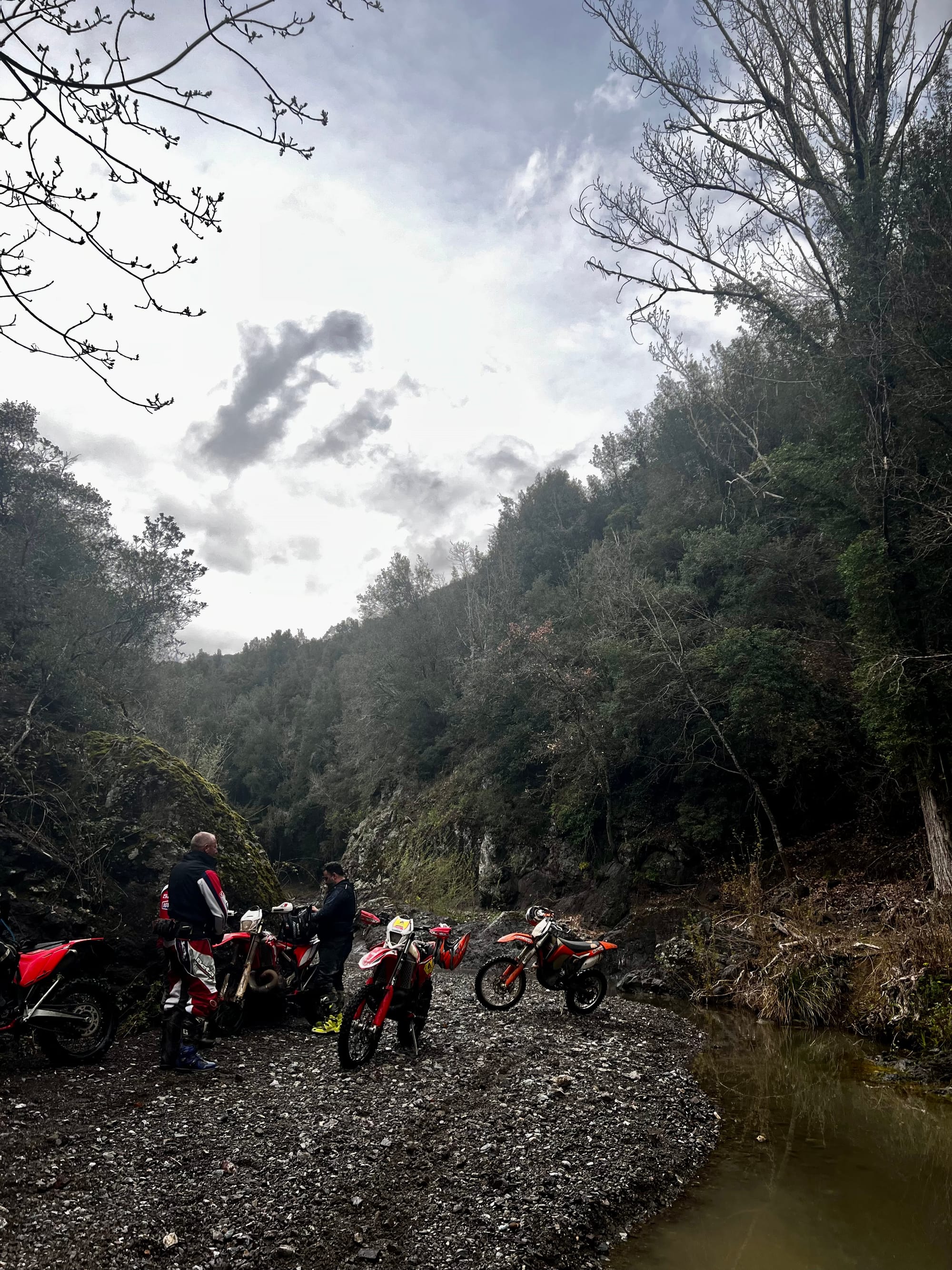 Das Foto zeigt drei Personen, die neben vier Enduro-Motorrädern an einem felsigen Ufer eines kleinen Flusses oder Baches stehen. Die Personen sind mit Motorradkleidung ausgestattet, darunter Helme und Schutzkleidung. Die Motorräder sind rot mit weissen Akzenten. Es ist ein bewölkter Tag, und die Umgebung wirkt natürlich und unberührt, mit kahlen Bäumen und Büschen im Hintergrund. Die Szene vermittelt ein Gefühl von Abenteuer und Outdoor-Freizeitaktivitäten.