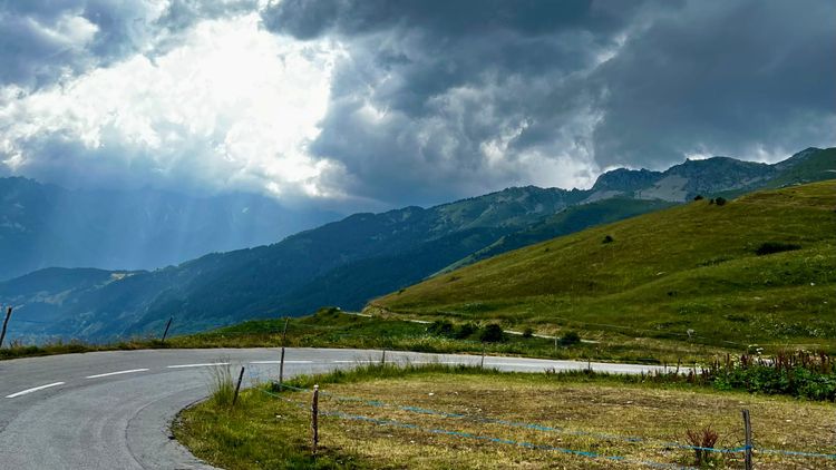 GPT  Alpine Szene mit kurviger Straße, Sonnenstrahlen durch Wolken, drohender Sturm.