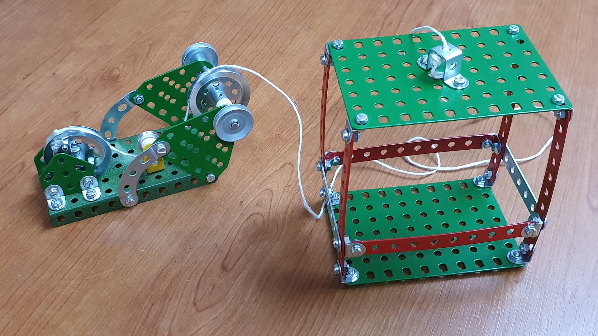 Two Merkur construction set models assembled from metal plates, screws, and pulleys on a wooden floor.