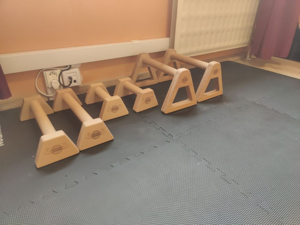Wooden parallettes arranged on a foam exercise mat next to a wall, used for calisthenics training in a home environment.