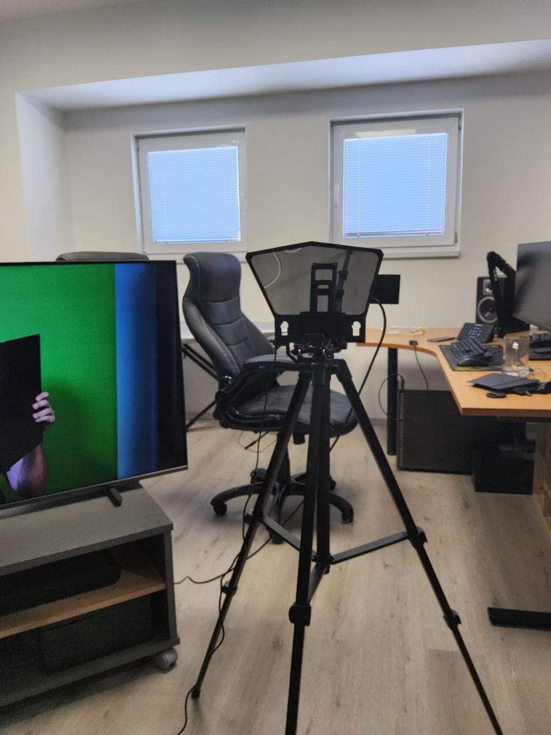 Improvised video recording setup in an office, with a teleprompter on a tripod, desk monitors, chairs, and work equipment arranged for regular speech practice.