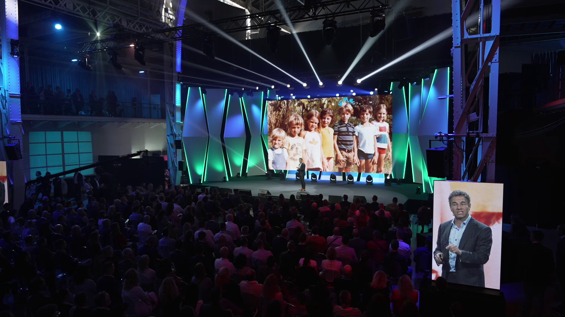 Wide shot of a large business conference stage with a speaker presenting to an audience, with a large screen behind him and a close-up video feed on the side.