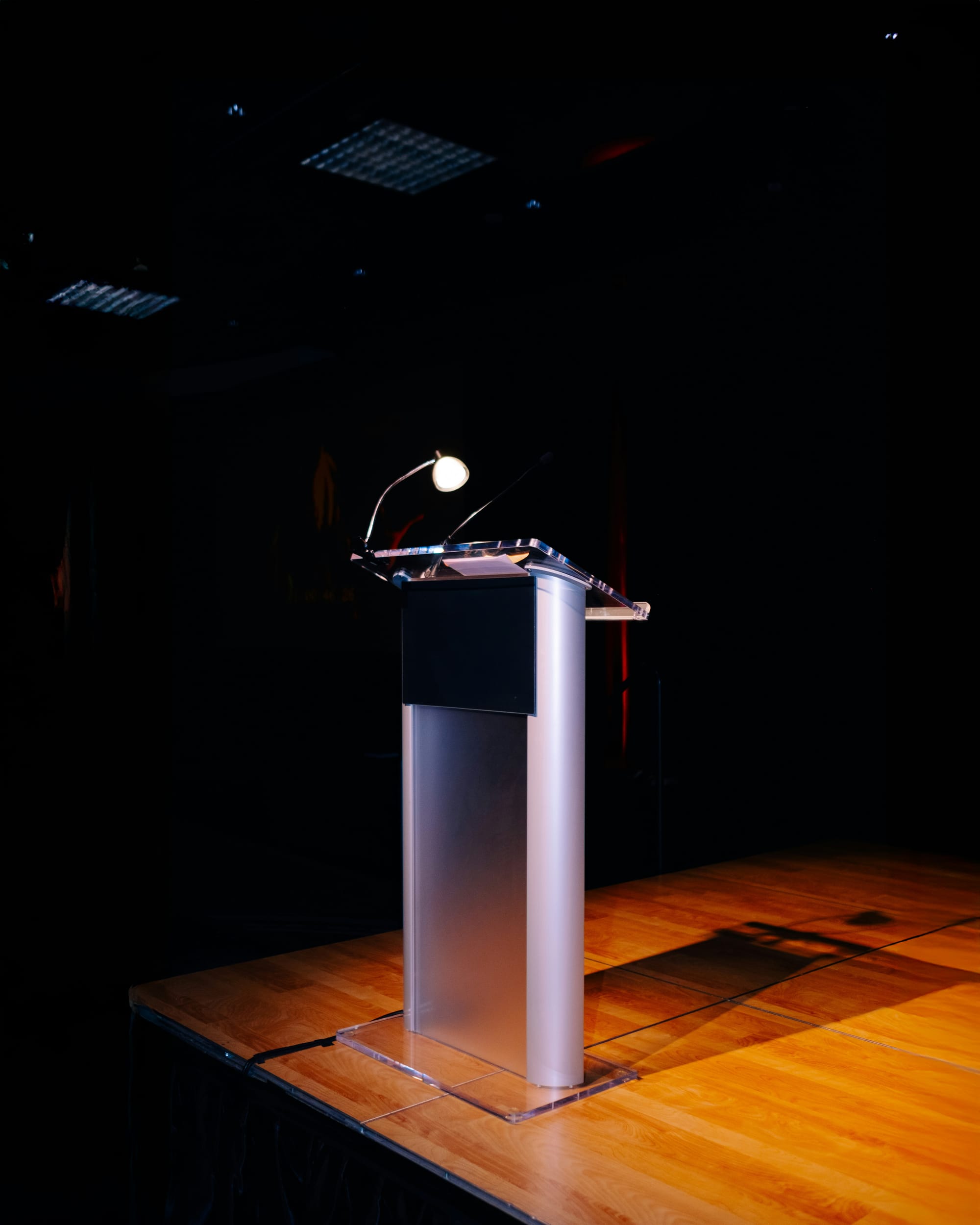 A single lectern with microphones and a small reading light stands on an empty wooden stage against a dark background, suggesting a formal speaking setup before a speaker arrives.