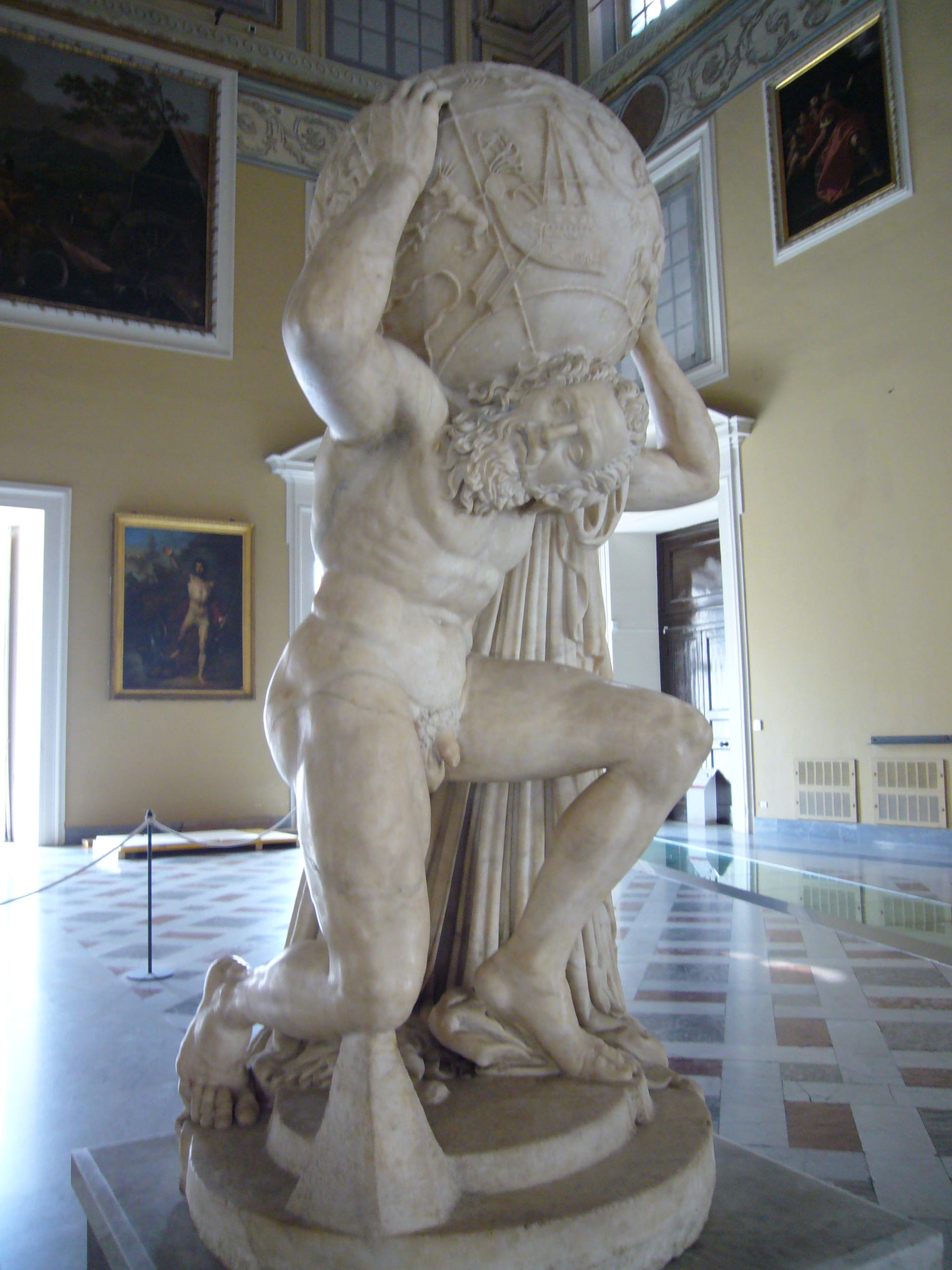 Marble statue of the Greek Titan Atlas kneeling under the weight of a large celestial globe, holding it above his shoulders with both arms.