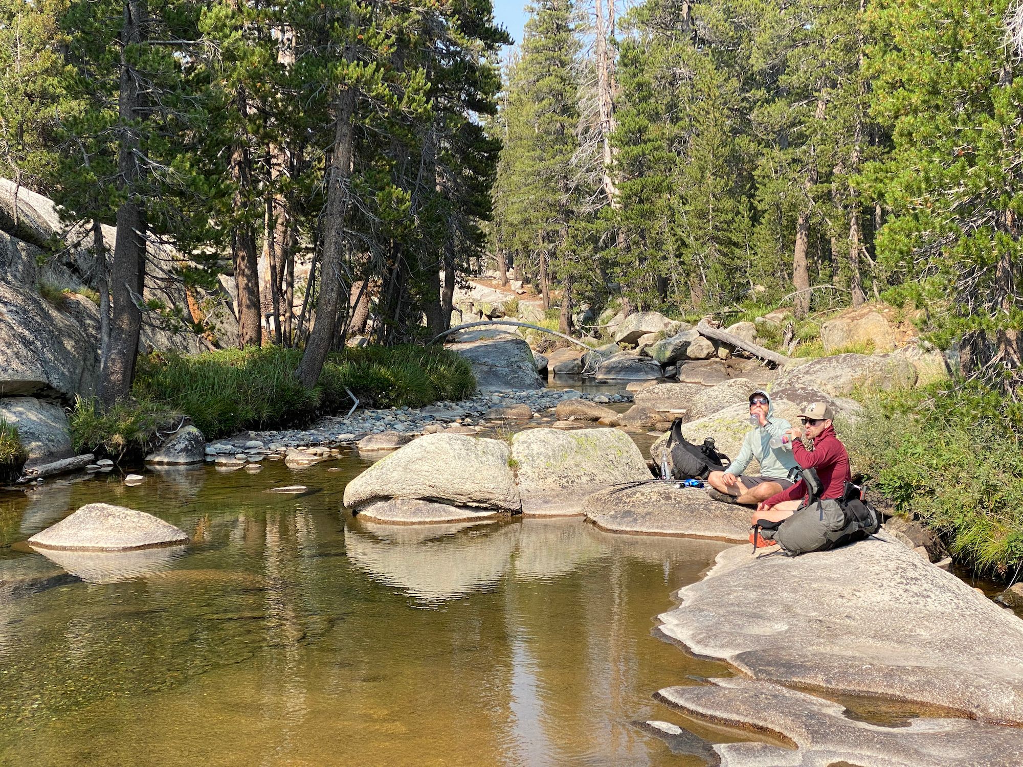 Two men sitting by a creek drinking water. 