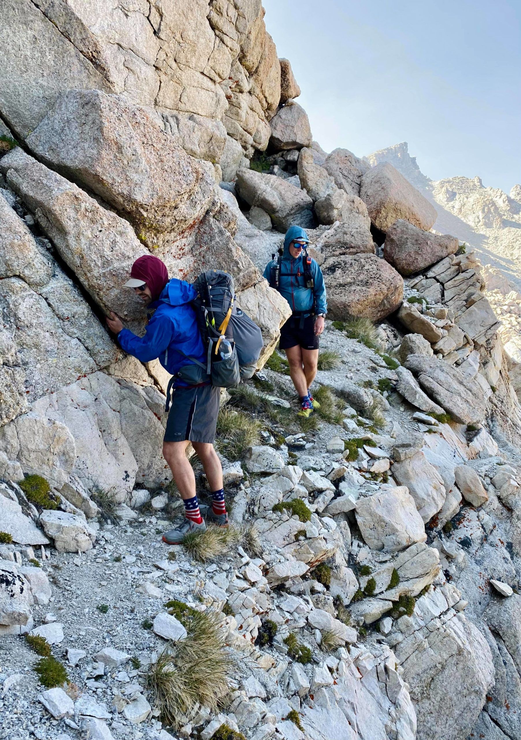 Two backpackers. The first is holding on to a rock wall.