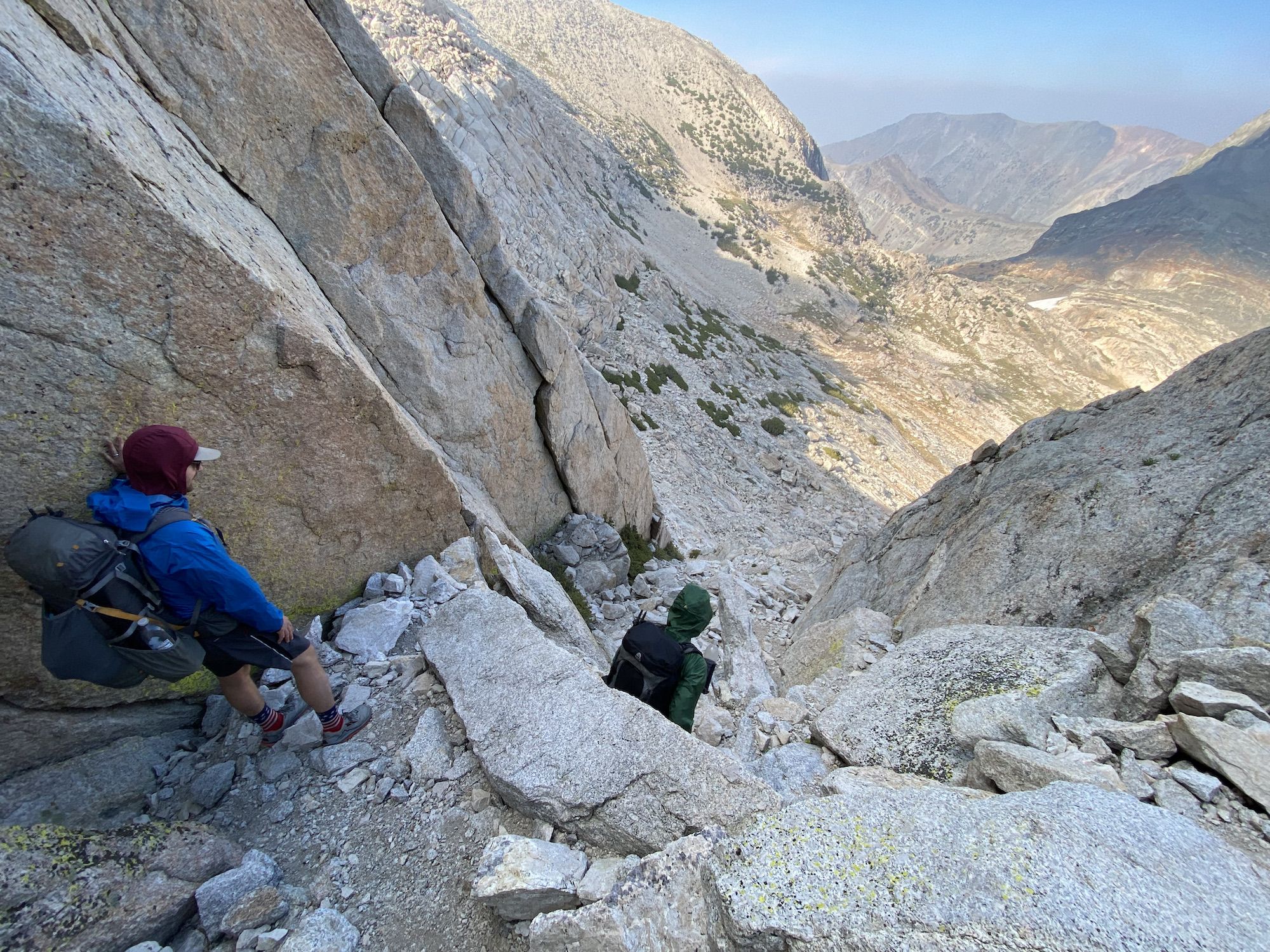 A backpacker standing below another backpacker under a large rock.
