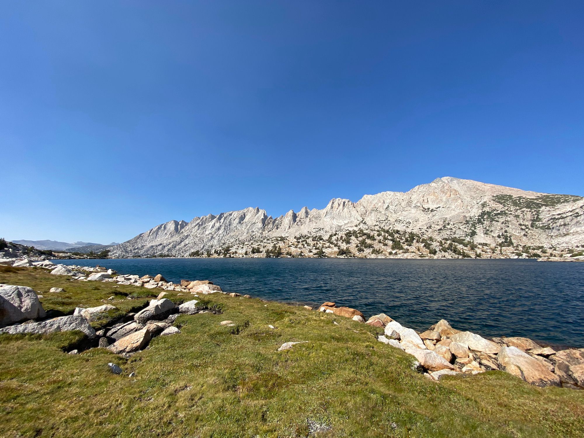 A grassy shoreline of a lake with jagged mountain peaks behind the lake.