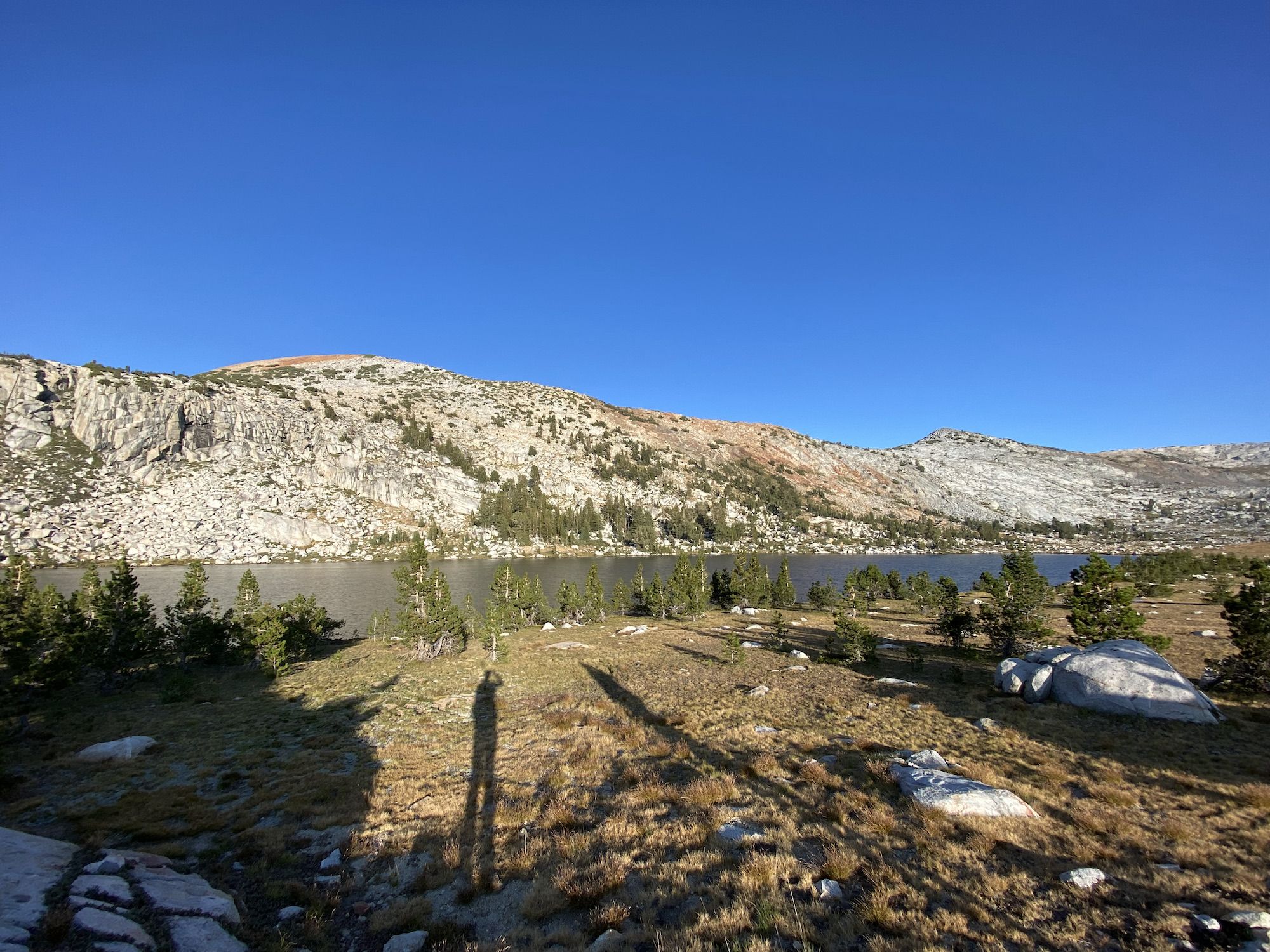 A lake with a meadow in the foreground. The long shadows of two backpackers can be seen.