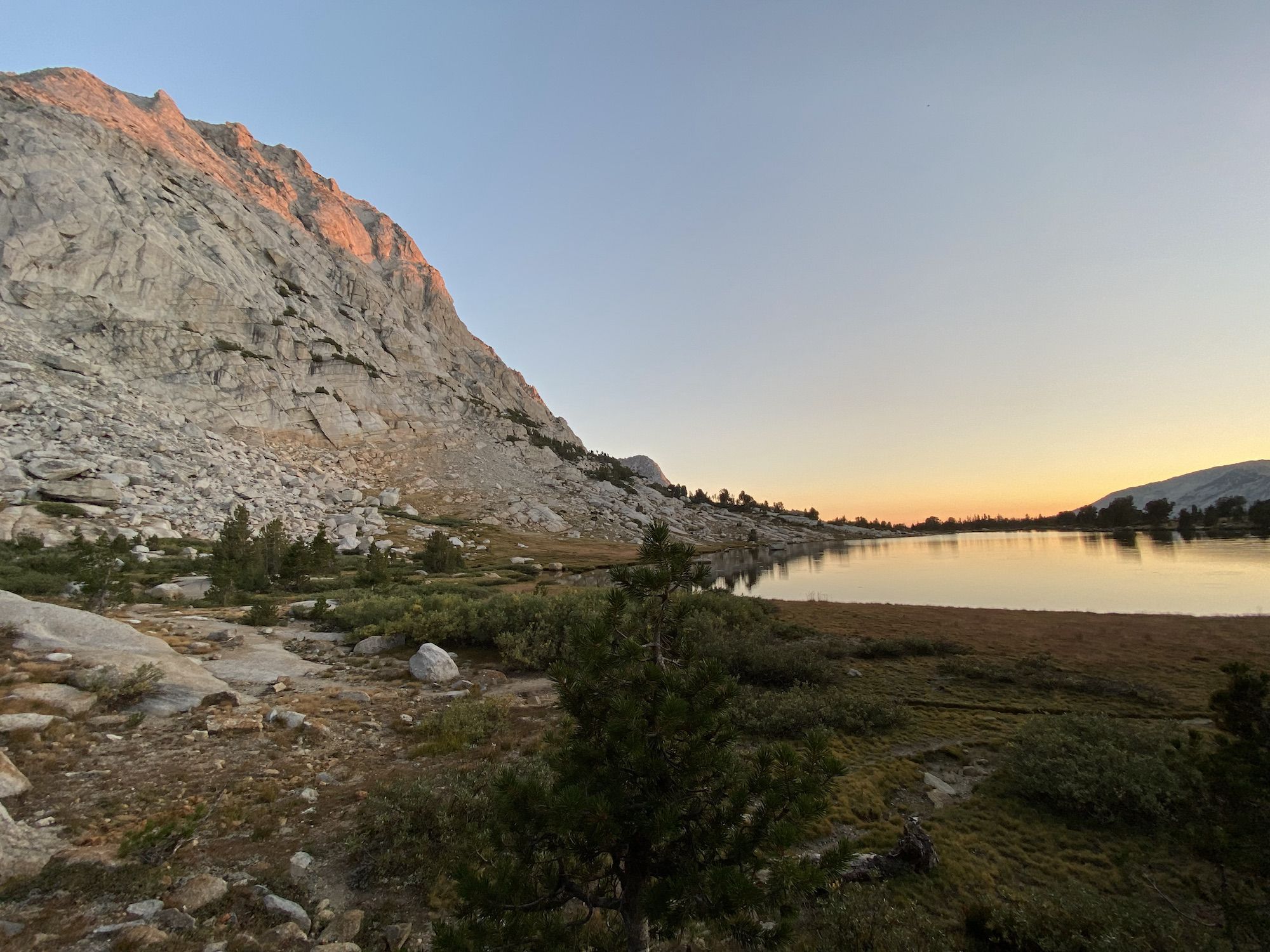 A calm lake with a large mountain on the right side, reflecting the orange light of the sunset.