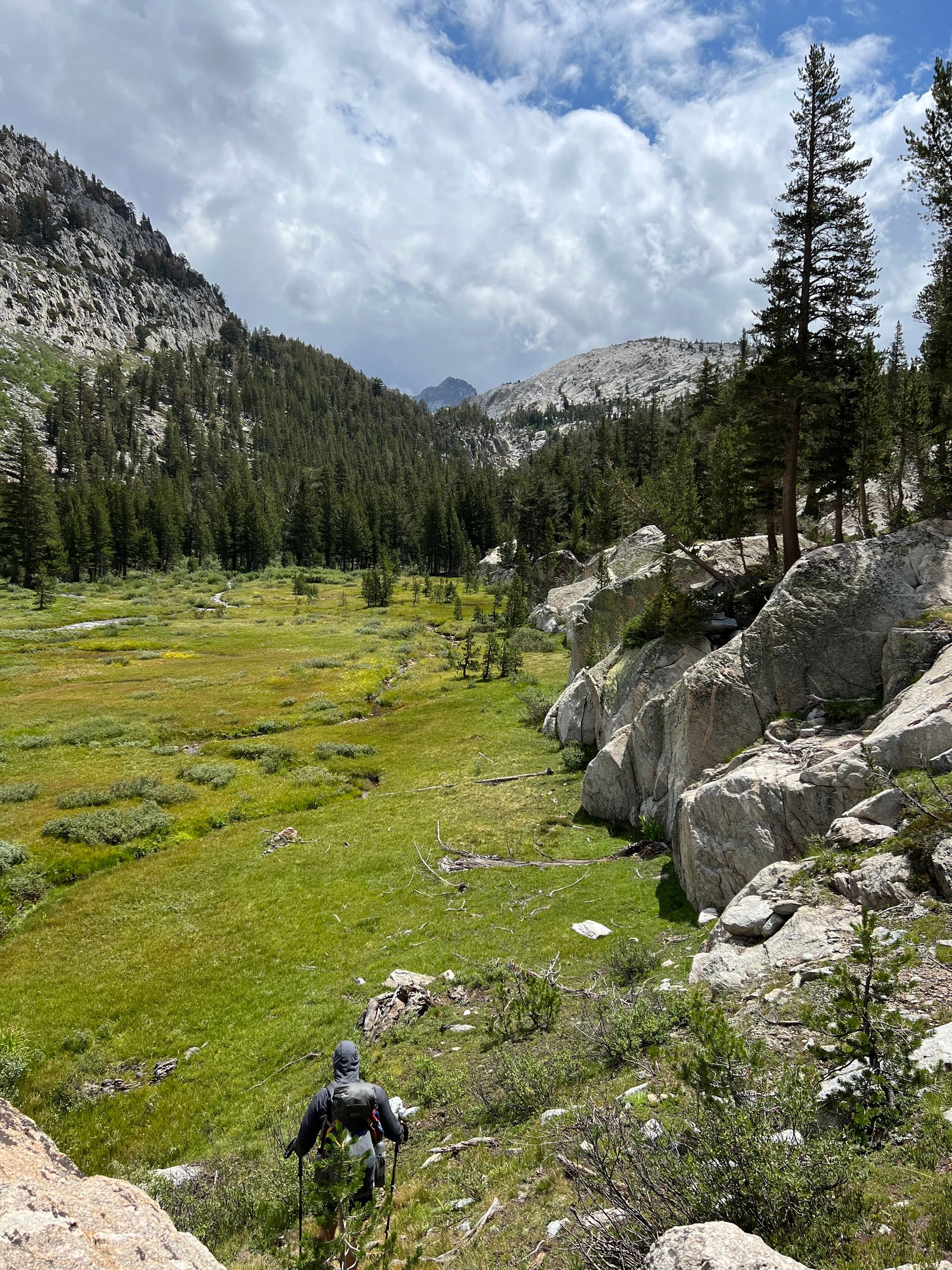 A backpacker walking onto a meadow. There are large granite boulders on his right side.