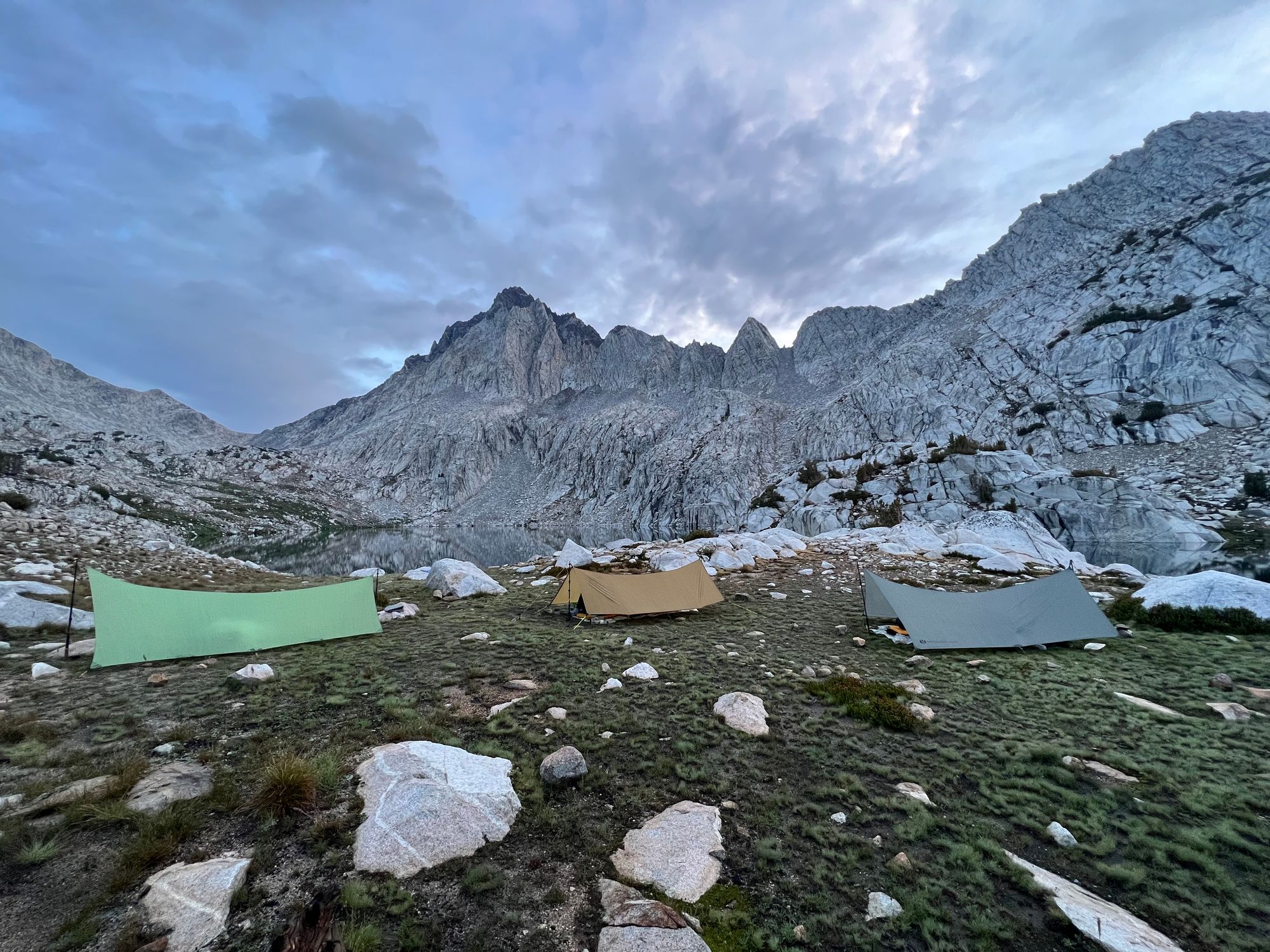 Three tarps in front of a lake. Large granite mountains on the far side of the lake.
