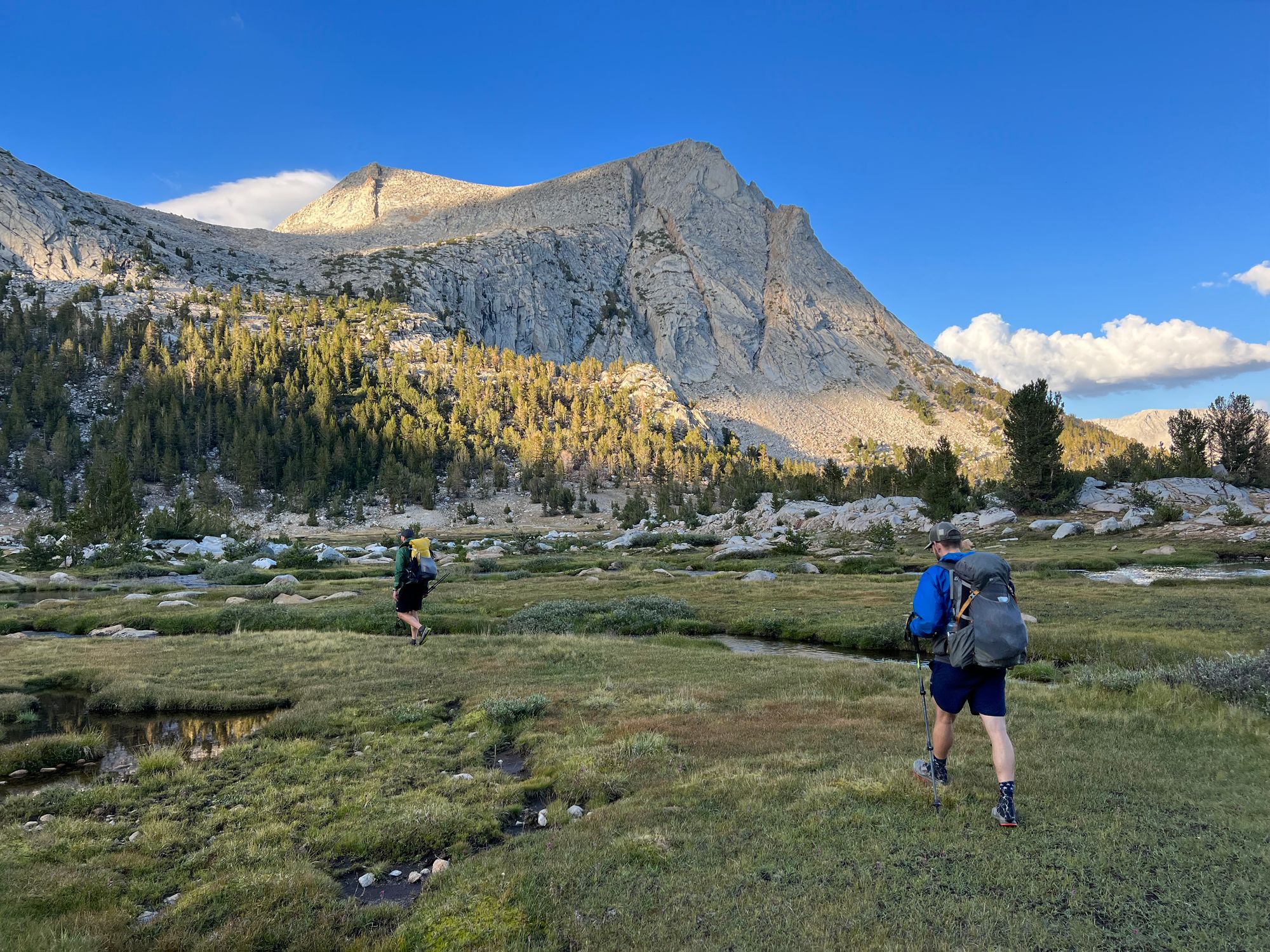 Two backpackers walking throug a meadow, along a stream.