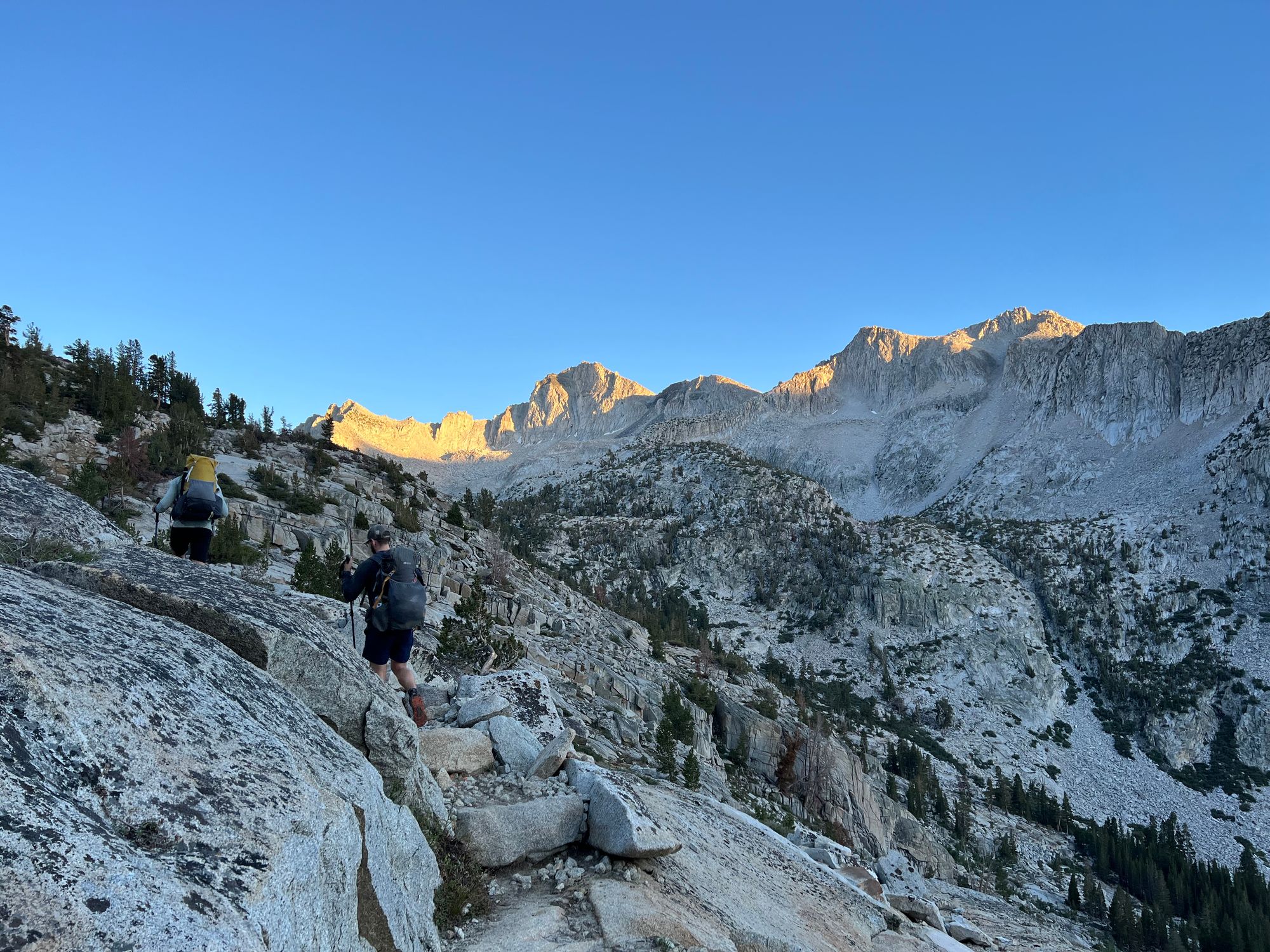 Two backpackers walking on a steep hillside. A wall of mountains in the distance.