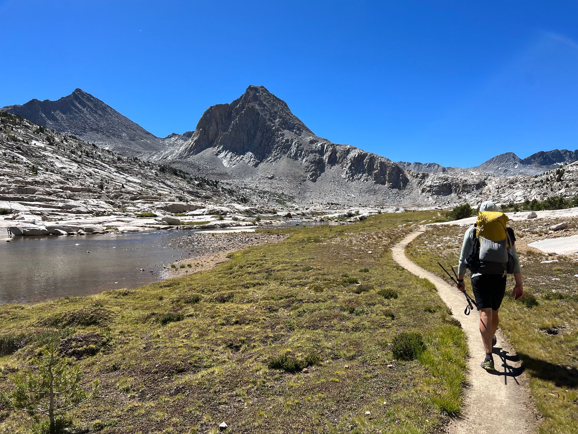 A backpacker walking along a path through grass, next to a creek. A large mountain in the distance.