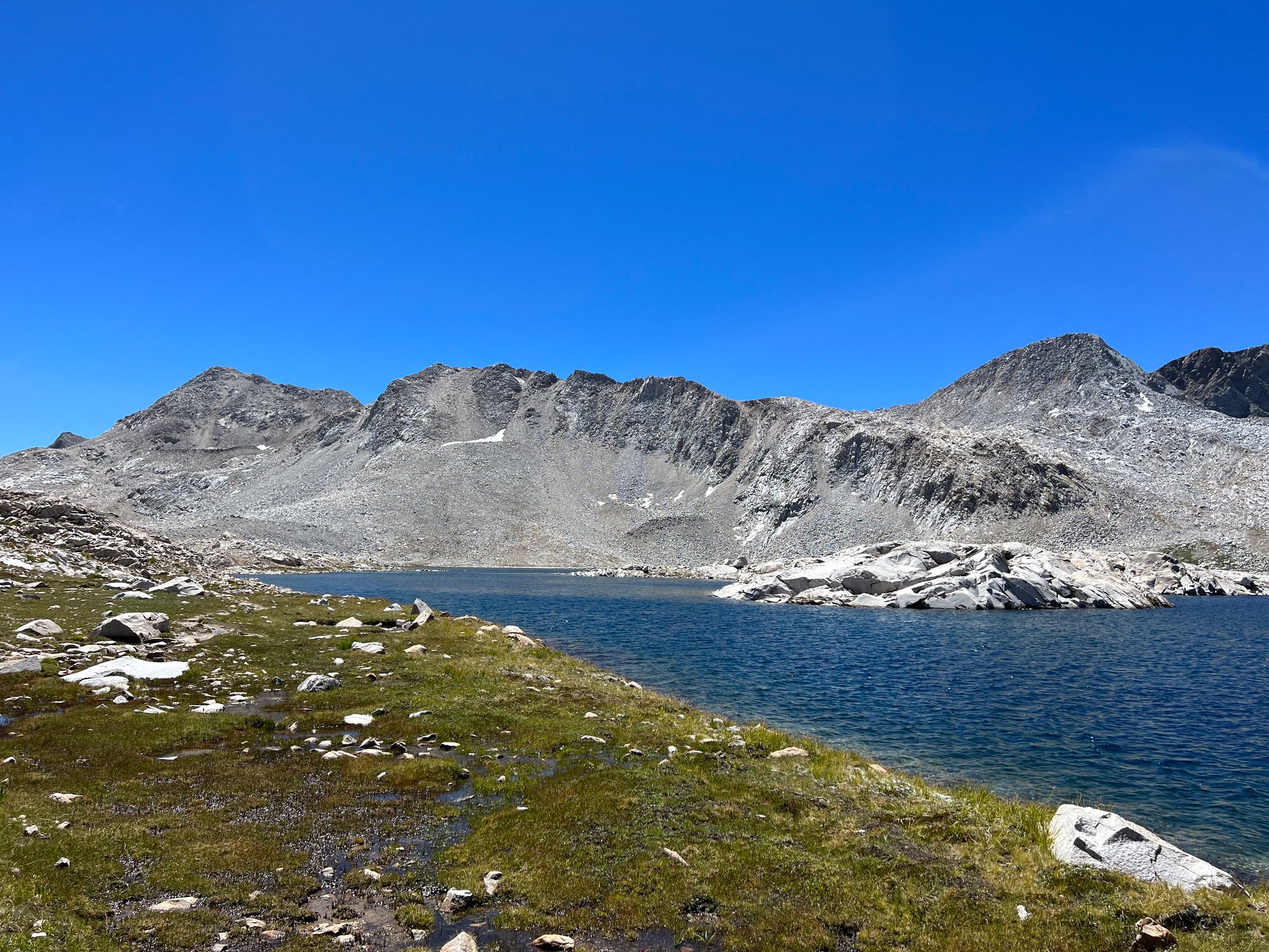 Marshy grass shoreline next to an alpine lake. A talus slope on the far side of the lake.