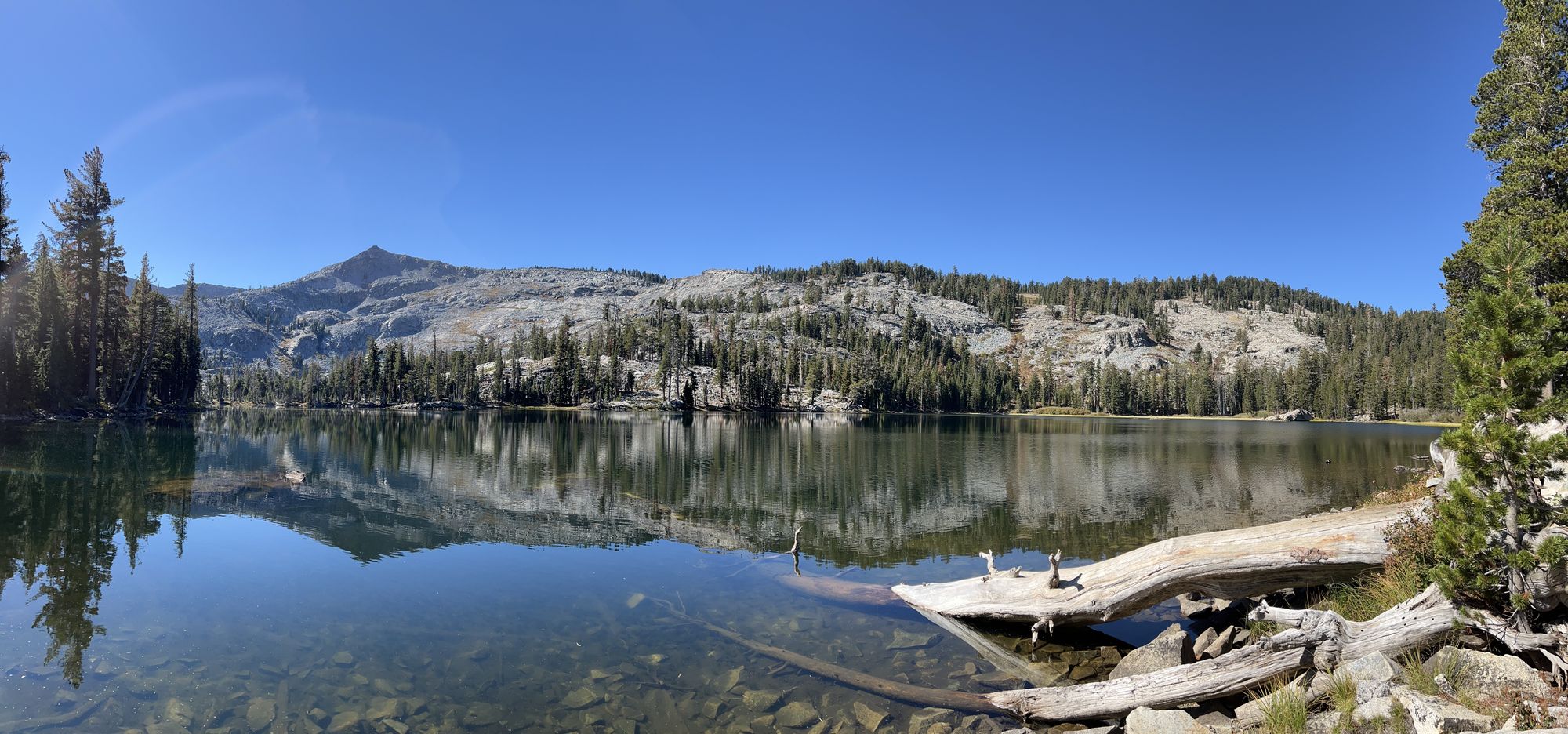 A still lake reflecting mountains in the distance. A fallen tree in the water nearby.