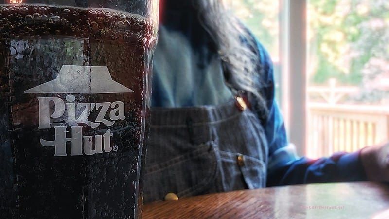 A clear plastic cup with the Pizza Hut logo, which contains root beer. The author is in the background.