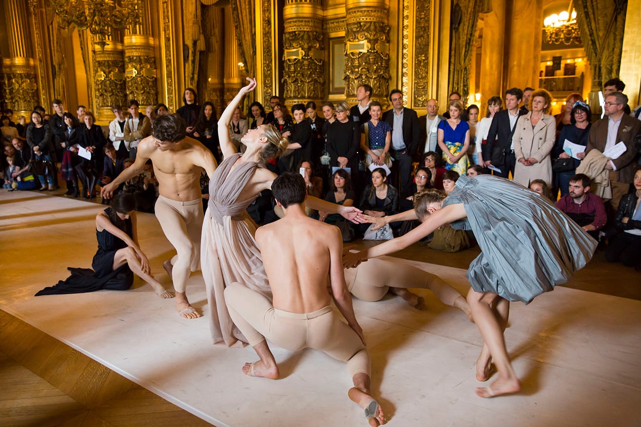 Six dancers, the men shirtless and the women in dresses, perform in an ornate ballroom for a well-dressed crowd of art world professionals and enthusiasts.