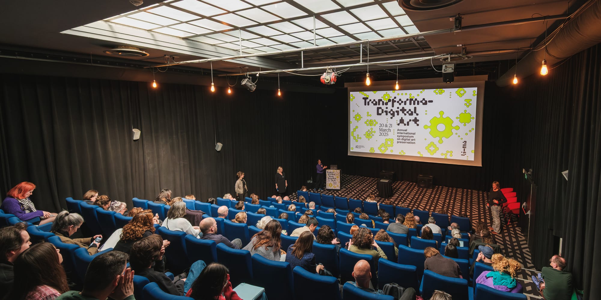 A screening room being used as a conference hall, with around 50 attendees sitting on plush blue seats and a few people on stage. The conference name Transformation Digital Art is projected on the screen.