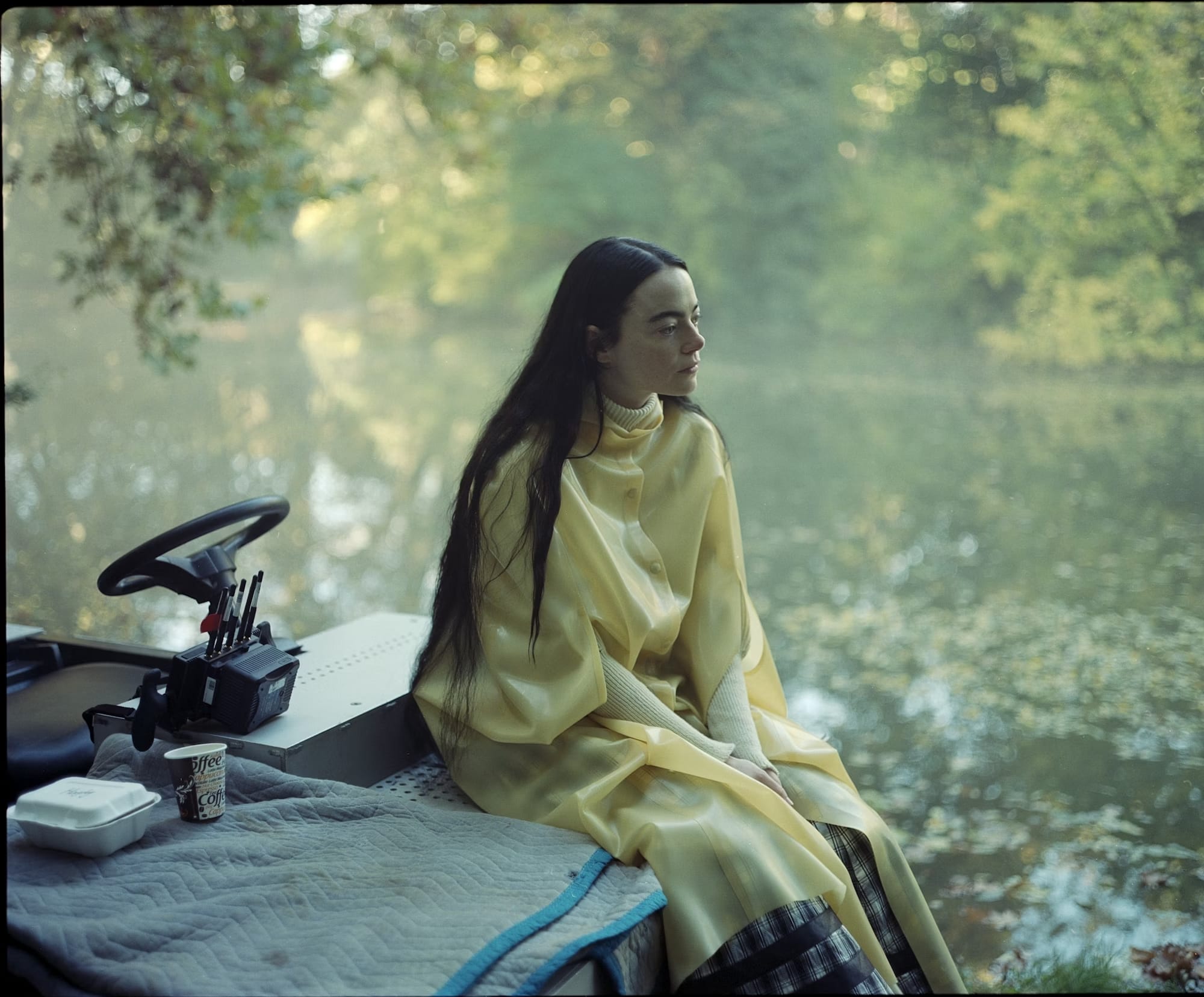 A young woman with long dark hair in a yellow coat sitting on the stern of a boat, with an out-of-focus pond covered in leaves behind her.