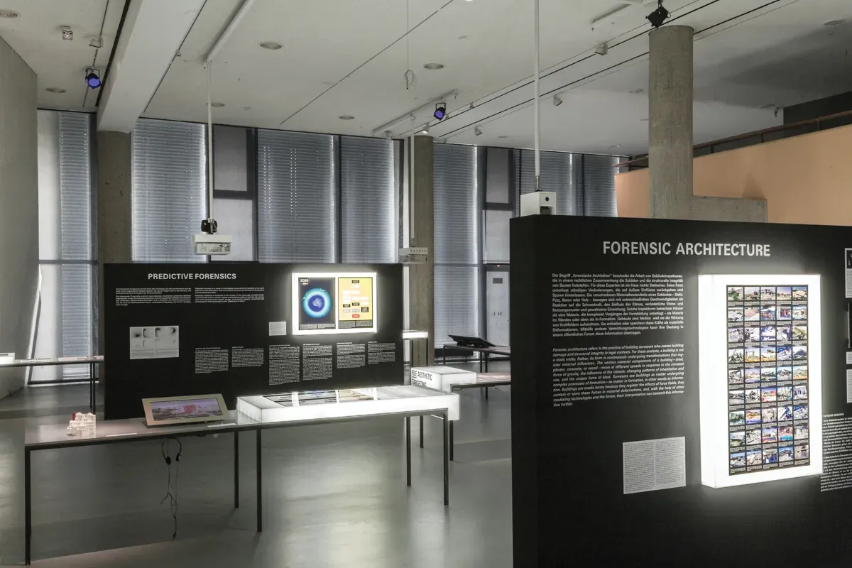 An installation, with two tables, each one with text and images posted on a black board, one has Predictive Forensics written on it and the other has Forensic Architecture written.