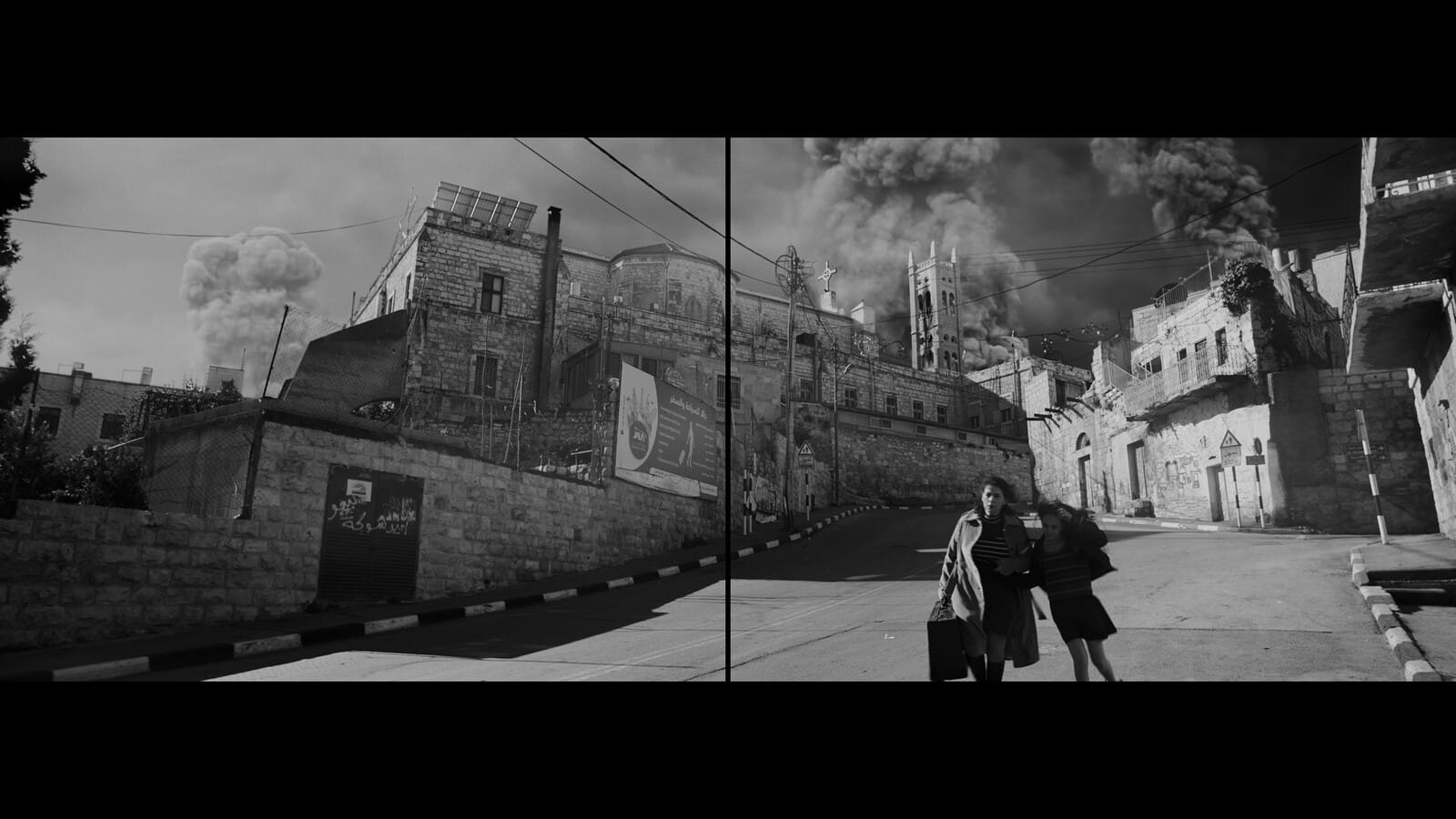 A woman with a suitcase and a young girl walking down a street with buildings engulfed in smoke behind them.