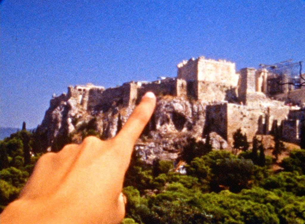 A grainy film frame with an outstretched hand, with a finger pointing at an ancient fortress in the distance.
