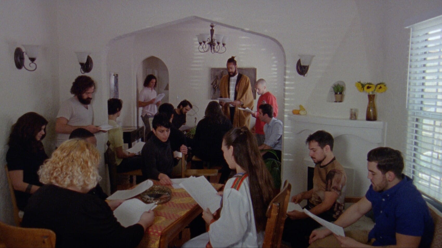 A large group of people inside a home, all holding and reading a few pieces of white paper.