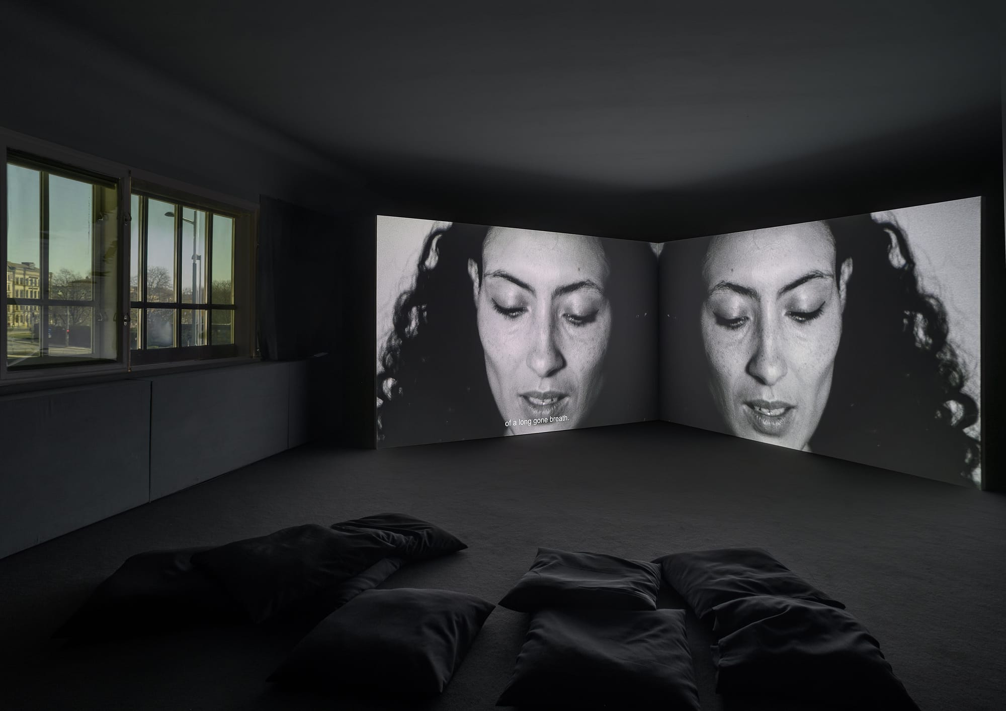 A dark gallery with black bean bags on the floor and two large screens with black and white images of a woman with dark curly hair.
