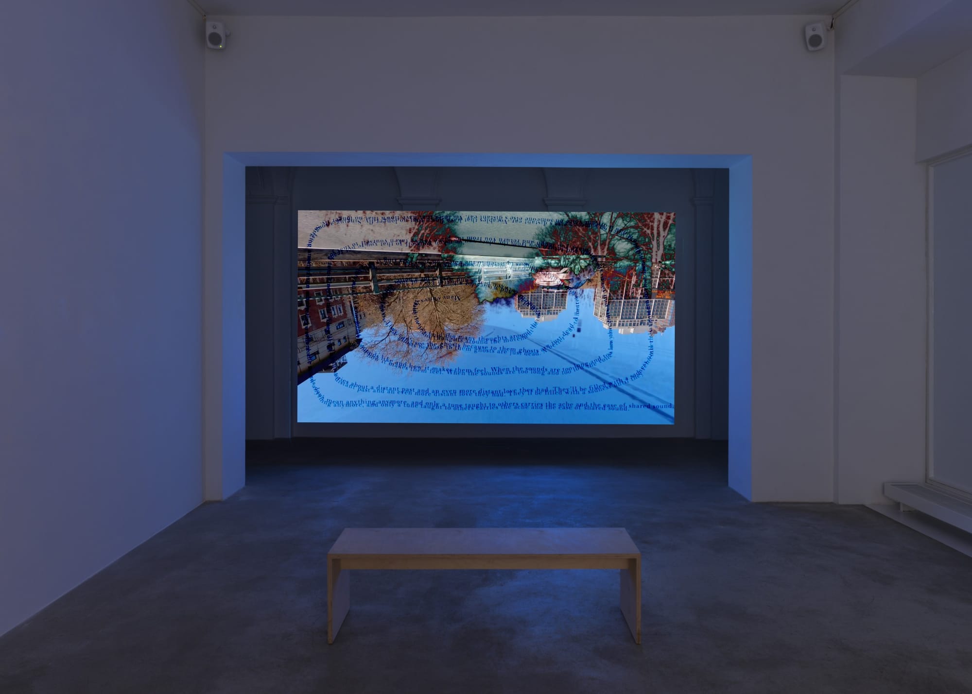 An installation view in a gallery, with a lone wooden bench and a video screen with an image of an upside-down landscape with poetry in the shape of a spiral printed on it.