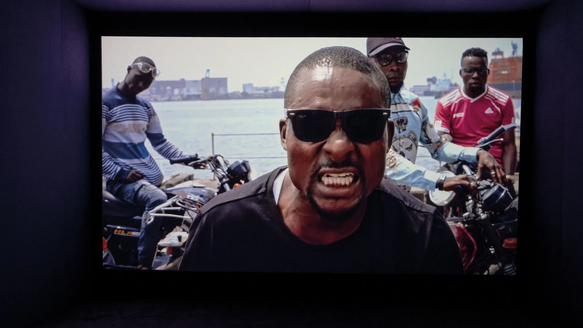 A Black man in sunglasses gritting his teeth and frowning at the camera, with men on motorcycles behind him.