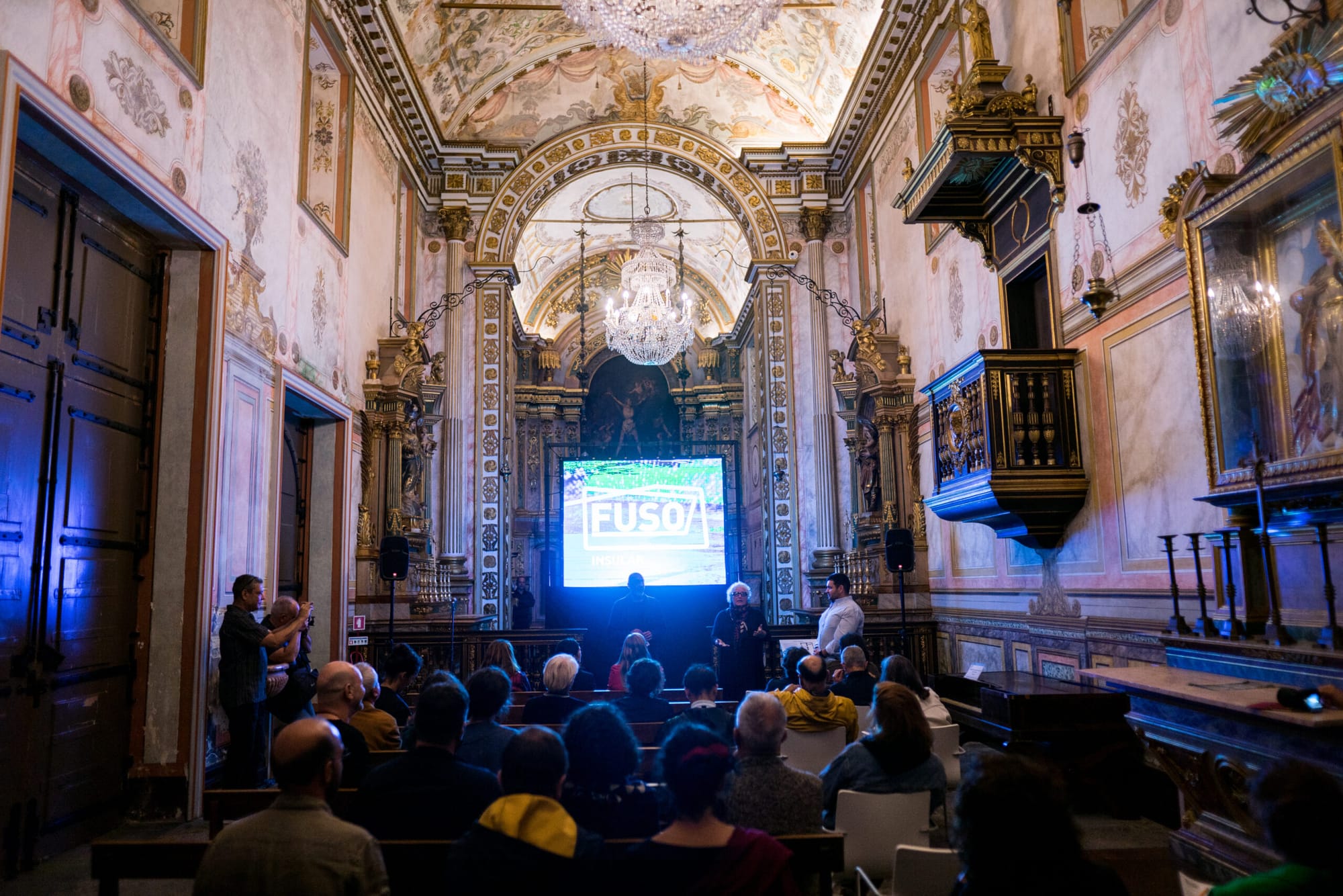 A screening in an ornate building with chandeliers and the graphic FUSO being projected on screen.