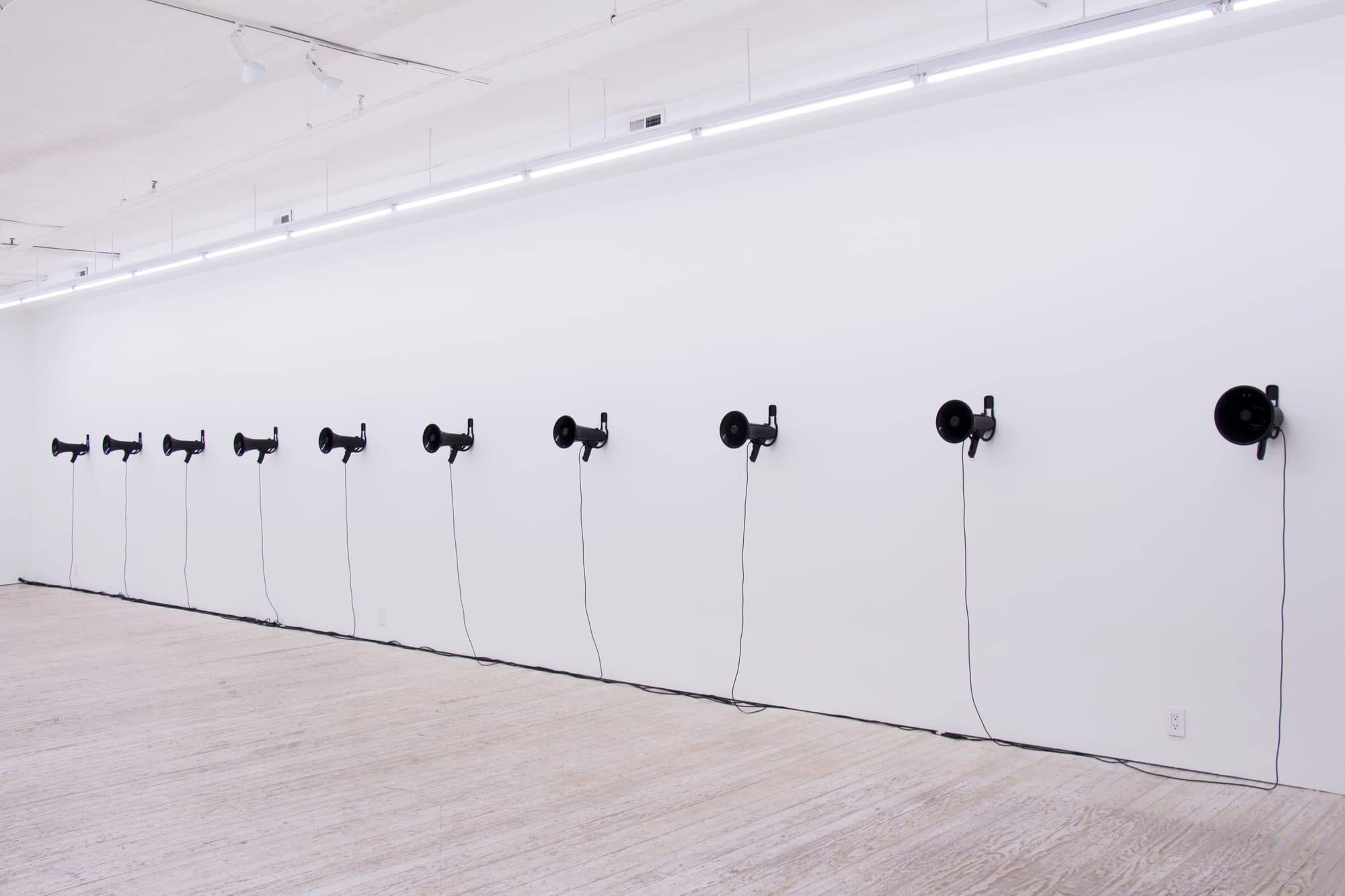 A white gallery wall with a row of ten black megaphones hanging on it.