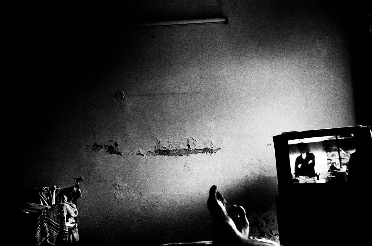 A black and white image of a cracked wall, with a television on the right side of the frame, and feet just visible at the bottom of the frame.