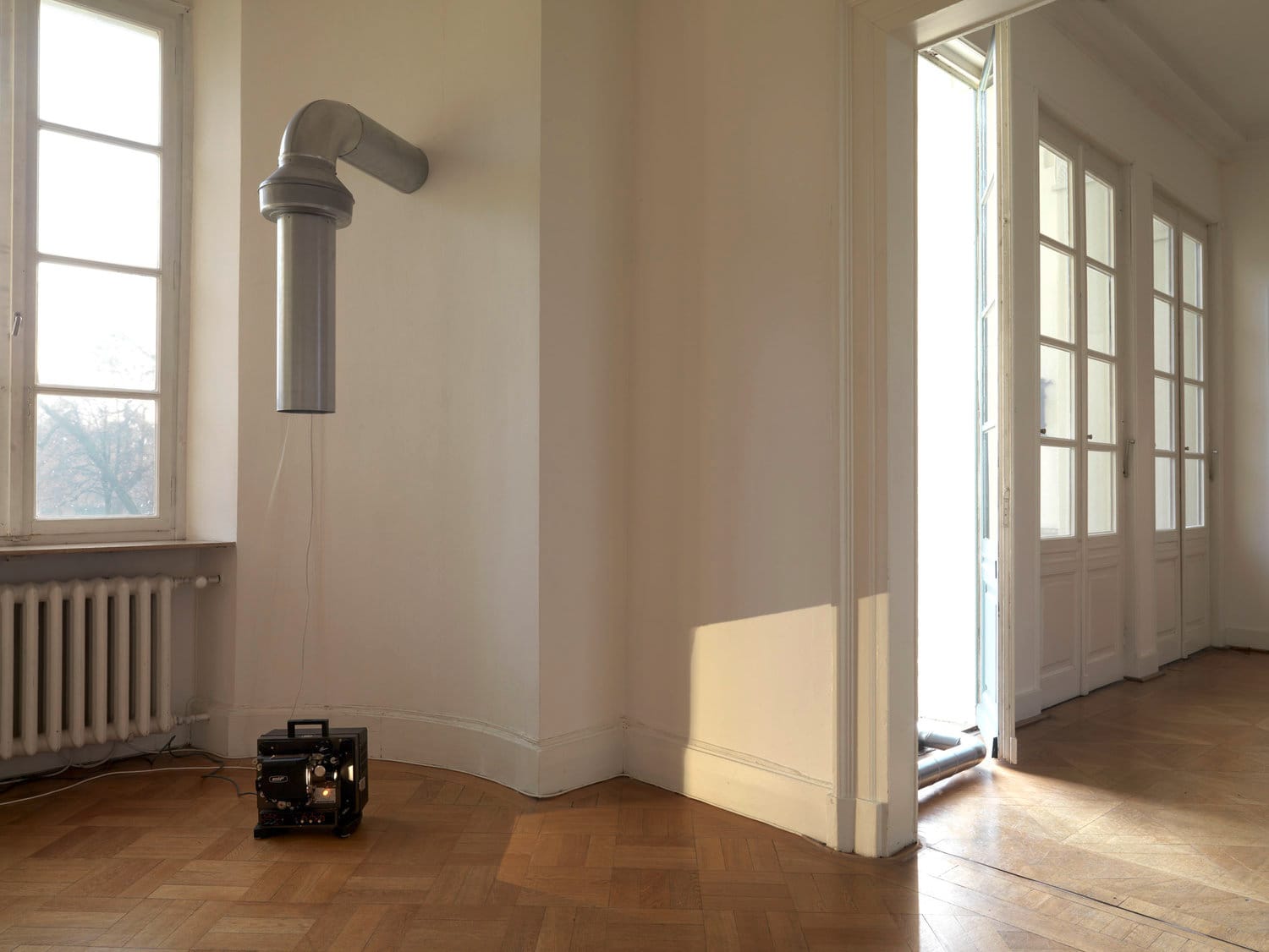 A sunlit gallery with hardwood floors, with a film projector on the floor and tubing above it that has a film strip coming out of it, with large windows in the background.