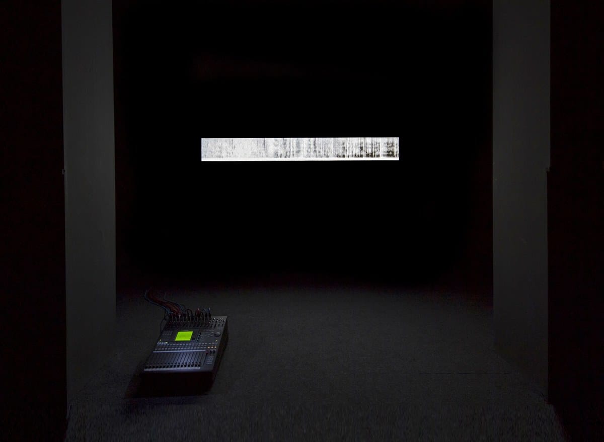 A darkened gallery with a mixing board on the floor and a small, horizontal projected image of an abstract diagram on the wall.