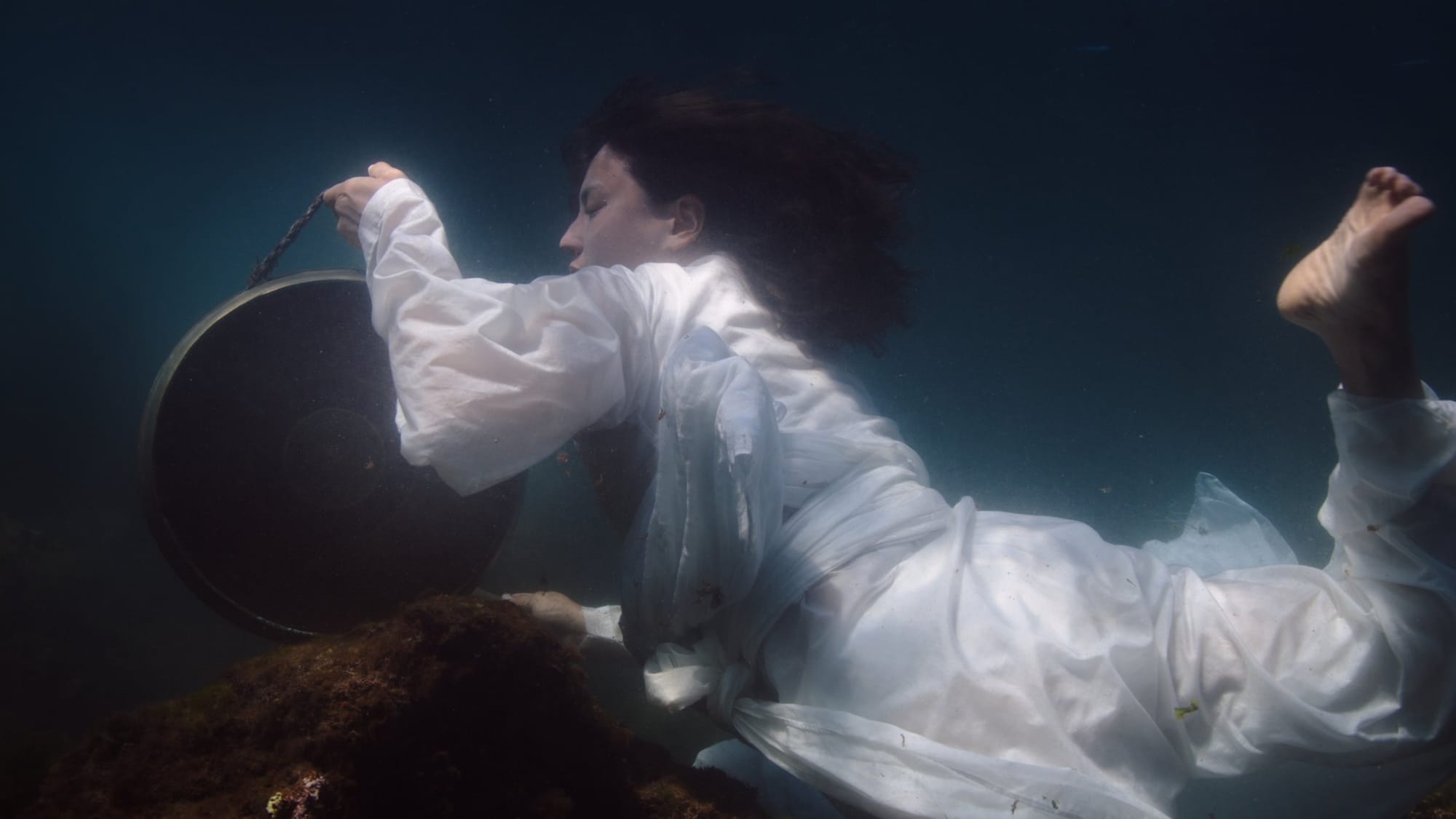 A woman dressed in white floating underwater and retrieving items from a wreck.
