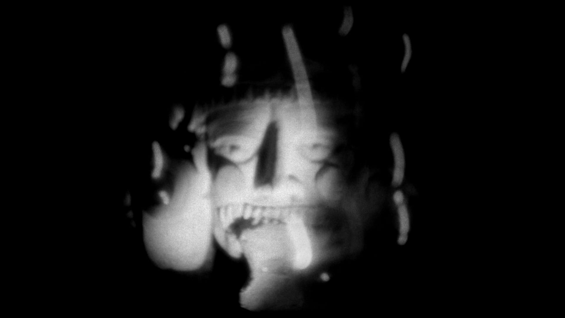 A grainy black and white film image of a ceremonial mask with grinning face.
