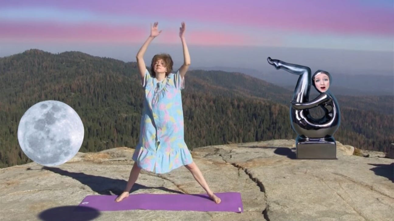 A woman in a colorful dress on an exercise mat on top of a mountain, with a full moon on the left and a sculpture with animated face on the right.