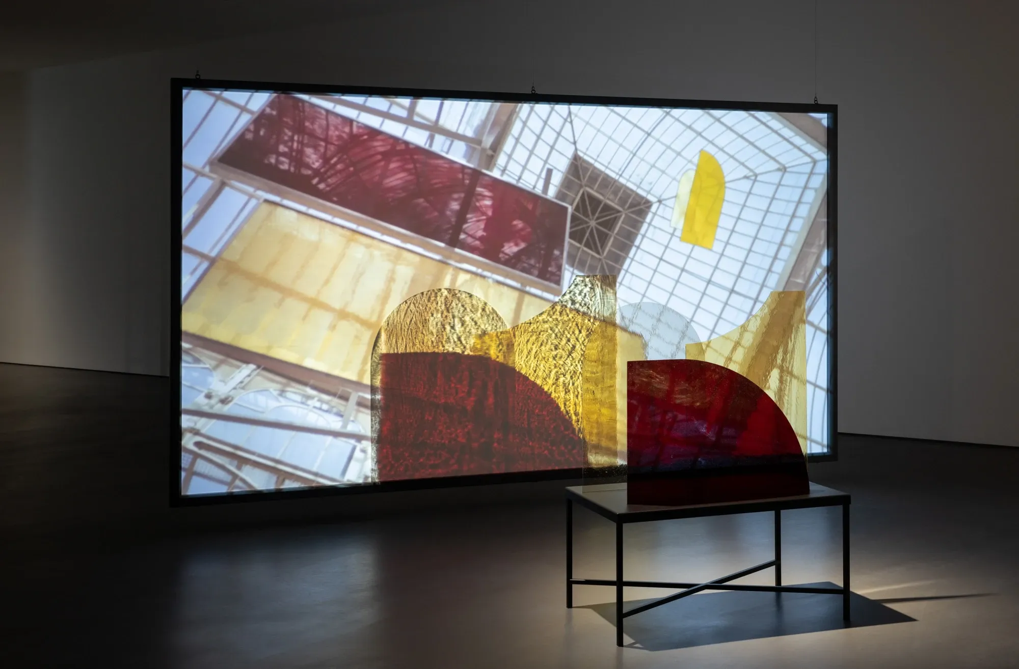An installation in a gallery with a large screen with colorful shapes projected onto it, and a small table in front of the screen with an abstract sculpture on it.