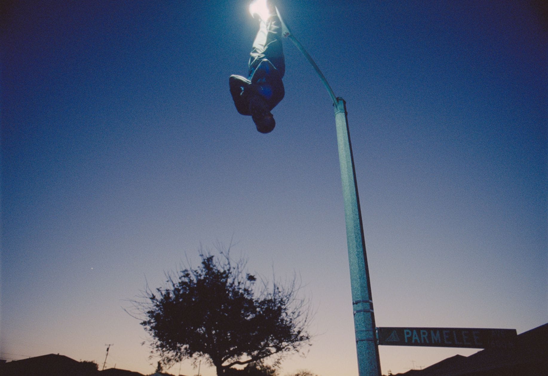 A man with his arms crossed hangs by his feet from a street light as dusk sets in.