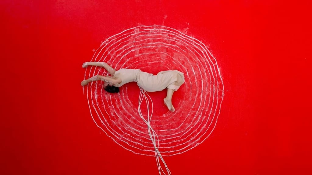 A high-angle view of a figure dressed in white lying down on a red floor with chalk circles drawn around their body.