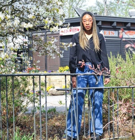 A Black woman with long hair wearing a black shirt and blue jeans stands behind a fence in a field of bushes and flowering trees.