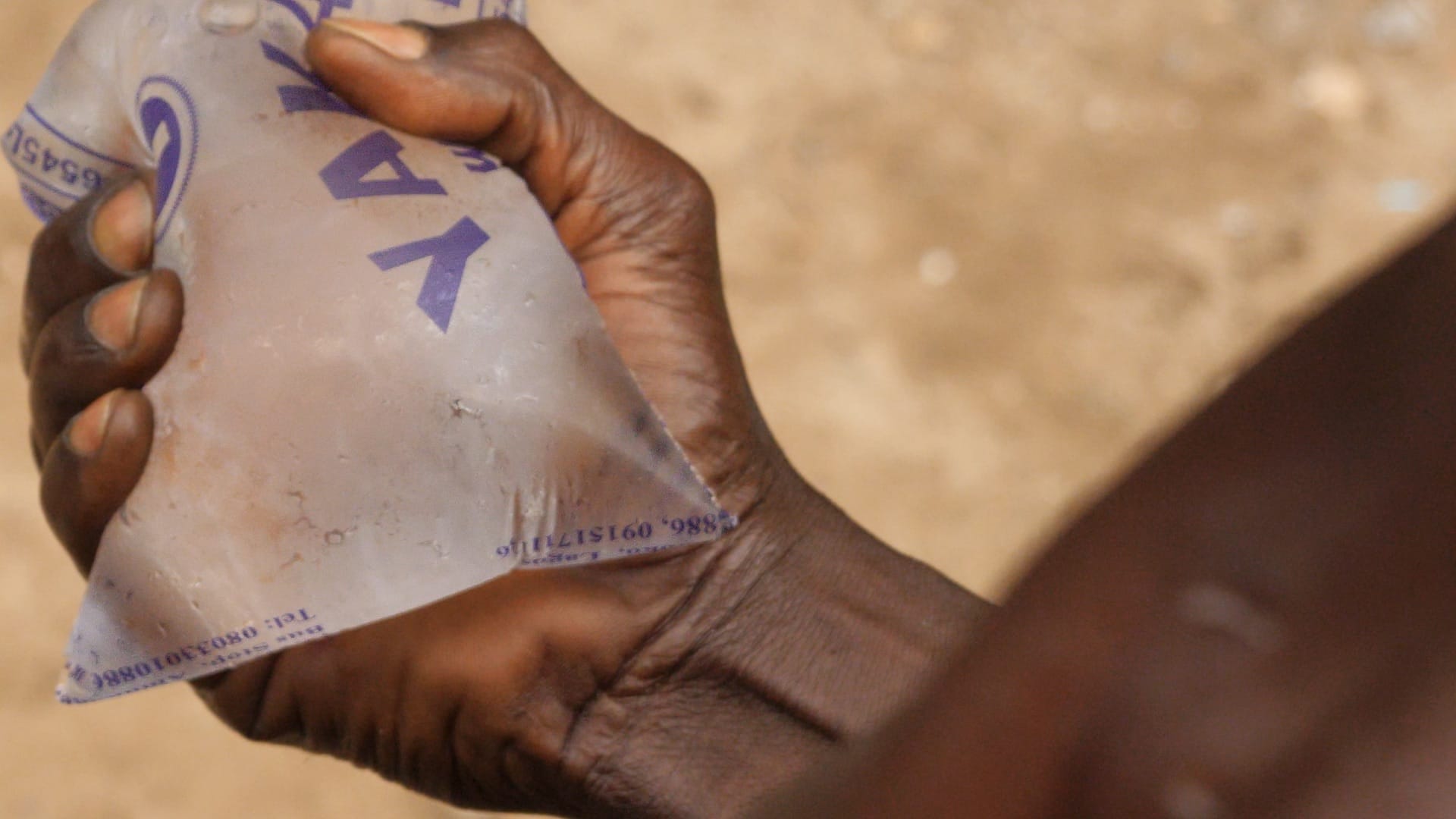 A close-up of a Black man's hand clutching a plastic bag of pure water.