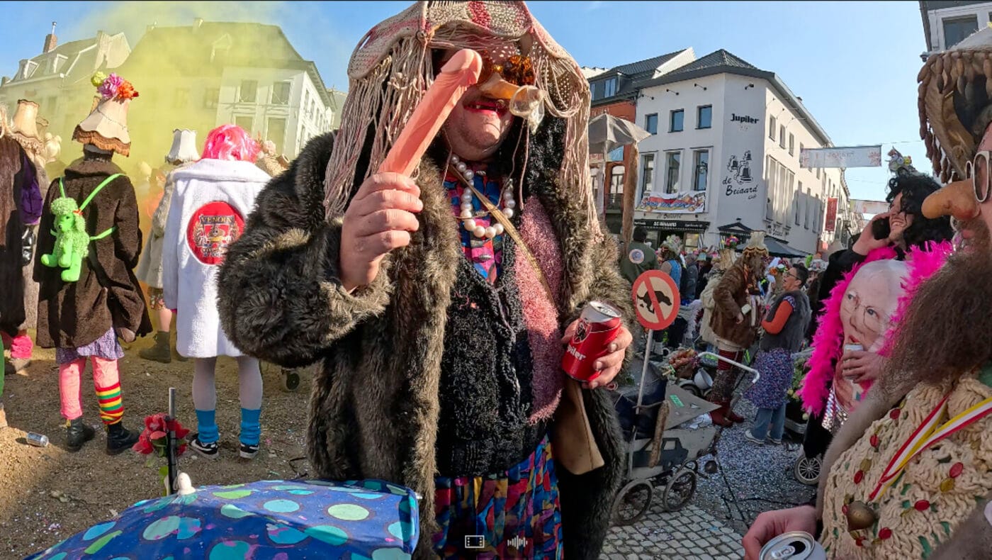 A man in an excessively surreal costume, holding a beer can and a prop penis, standing in the middle of a festival with people in wild attire.