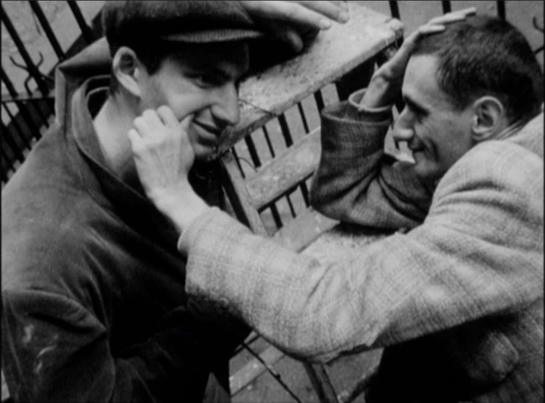A black and white archival image with two men smiling at each other, one pinching the other's cheek.